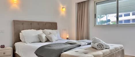 A bedroom with a bed, bedside tables, a window, and a view of buildings outside.