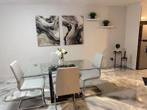 Ein moderner Essbereich mit einem Glastisch, umgeben von weißen Stühlen, zwei abstrakten Gemälden an der Wand und einem kleinen Blumentopf auf einem Regal.
