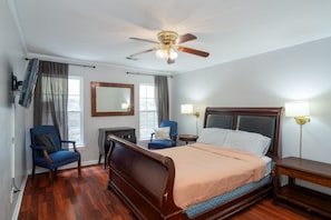 Ein Hotelzimmer mit einem hölzernen Bett, einem Schreibtisch, einem Stuhl, einem Deckenventilator und einem großen Fenster mit Vorhängen.