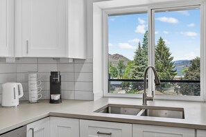 Una cocina con armarios blancos, una cafetera y vistas a una montaña y árboles a través de una ventana.
