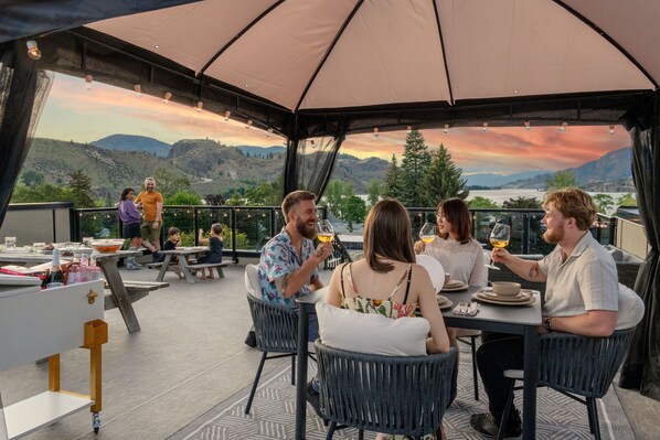 Un groupe de personnes est assis à une table en plein air, profitant d’une vue panoramique sur les collines et d’un coucher de soleil.