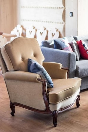 Un fauteuil capitonné avec une structure en bois, placé dans une pièce dont le sol est en bois et où l’on aperçoit, à l’arrière-plan, un canapé gris.