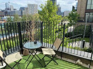 A balcony with a glass table and chairs, surrounded by a metal fence, overlooking a cityscape with buildings and greenery. - Spacious 2-bed Canal-side Apartment Sleeps 4 (Salford)