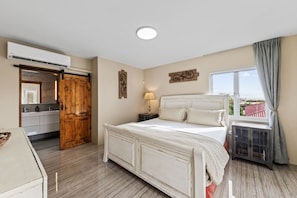 Een slaapkamer met een groot bed, een nachtkastje, een lamp, een raam met gordijnen en een schuifdeur naar een badkamer.