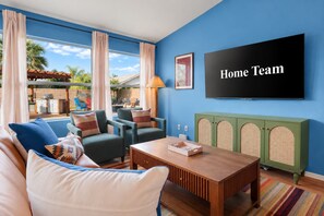 A living room with a blue wall, a flat-screen TV, a wooden coffee table, and a green cabinet with a rattan design. - The Sunny Social | Pool • Hot Tub • Games Galore! (Chandler)