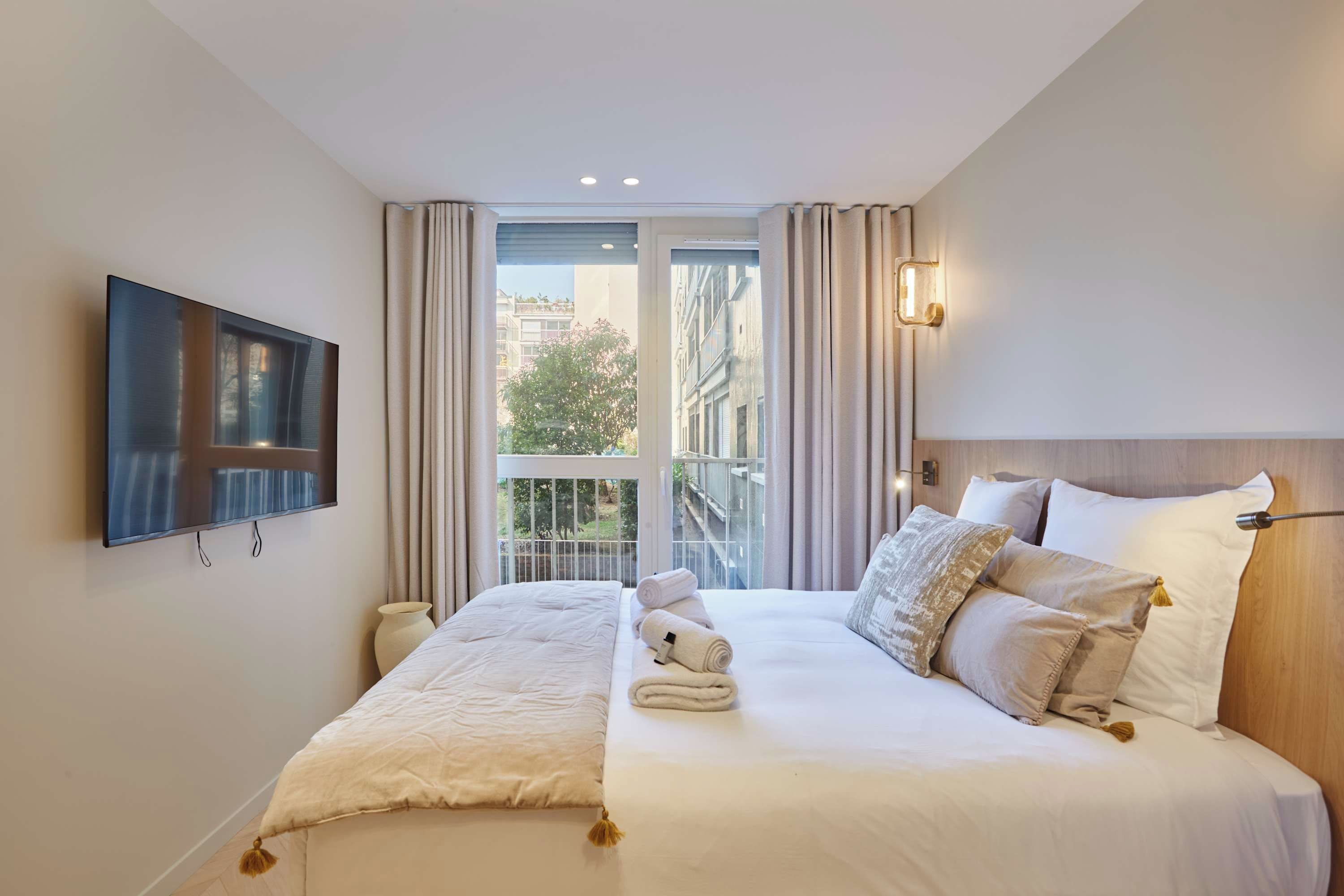 A hotel room with a bed, a television, and a view of buildings through the window.