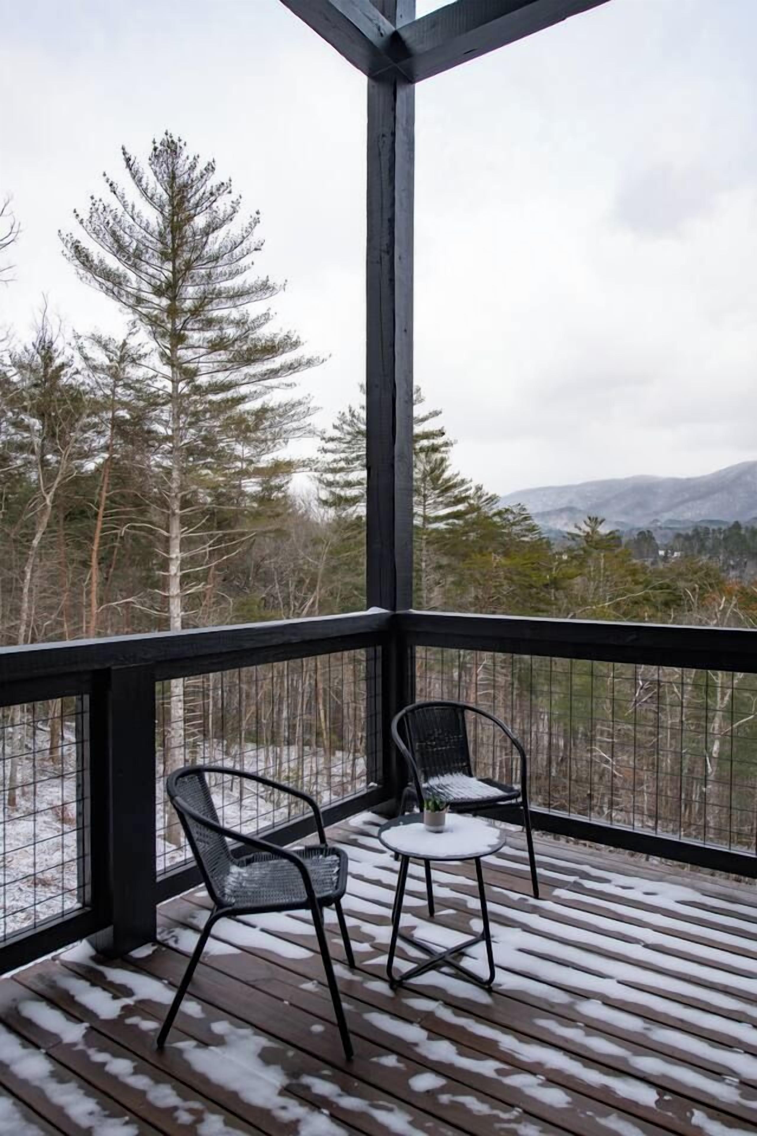 Um deck de madeira com duas cadeiras e uma mesa pequena, com vista para uma paisagem nevada com árvores e montanhas ao longe.