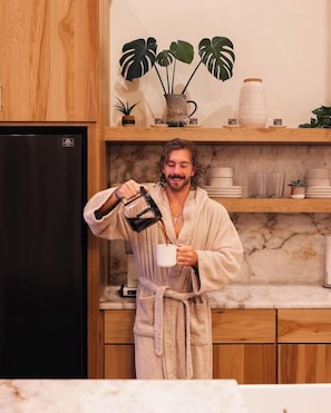 A person in a robe pouring a drink into a mug in a kitchen with wooden cabinets and a marble countertop.