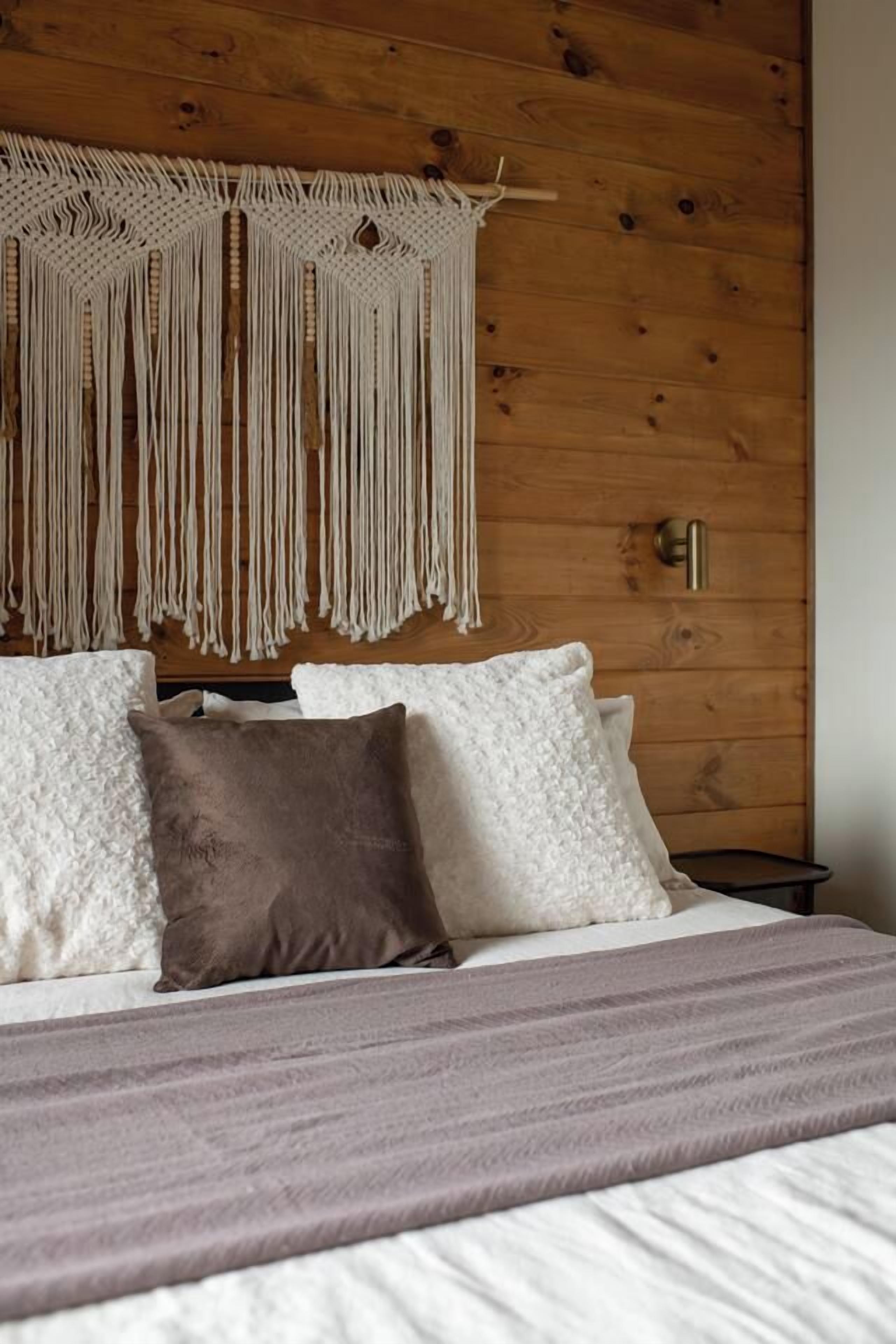 Uma cama com travesseiros brancos e castanhos, um pendente de macramé na parede e uma parede de madeira.