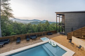 A modern outdoor pool area with wooden deck, lounge chairs, and a hot tub, overlooking a forested mountain range.