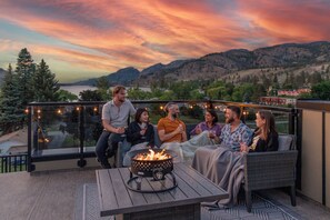 Un groupe de personnes est assis sur des meubles d’extérieur sur un balcon, près d’un foyer, et profite d’une vue panoramique sur un paysage magnifique, avec des montagnes et un lac.