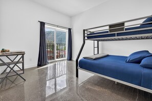 Uma cama beliche com colchão e almofadas azuis, uma escrivaninha pequena com um livro, e uma janela grande com cortinas.