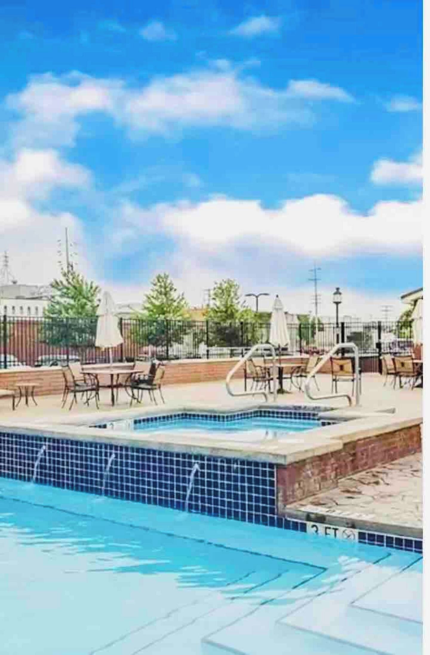Piscina com espreguiçadeiras e guarda-sóis, cercada por uma grade e árvores.