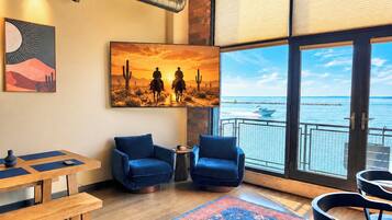A room with a large window overlooking a body of water, a painting of a sunset, and a wooden table with chairs.