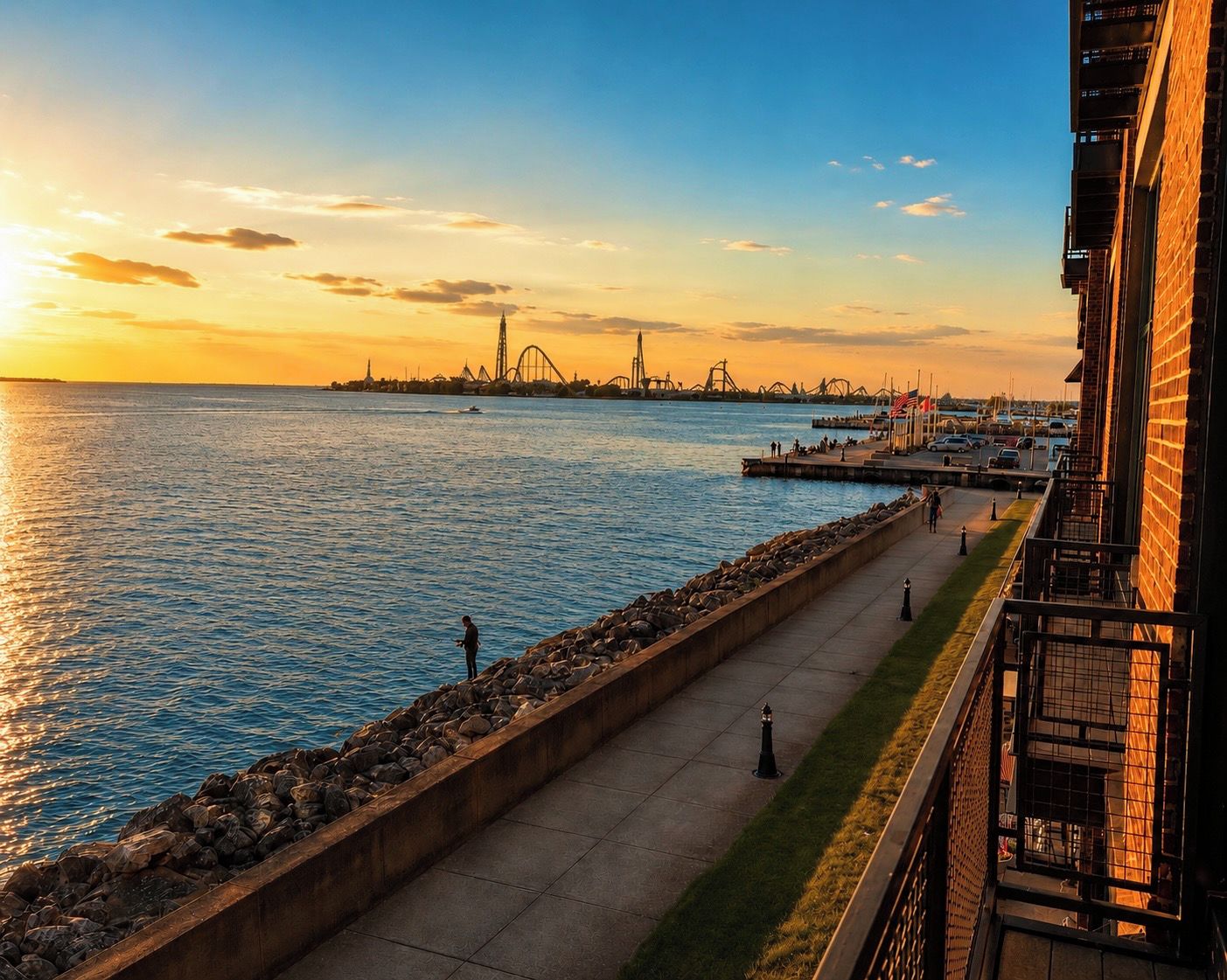 A waterfront promenade with a view of a distant roller coaster and a body of water reflecting the sunset.
