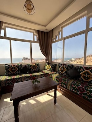 Een kamer met een zithoek, een houten tafel en grote ramen met uitzicht op het strand.