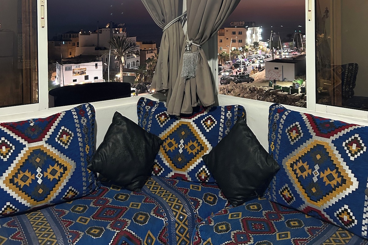A room with a large window, a patterned sofa, and a view of a cityscape at dusk.