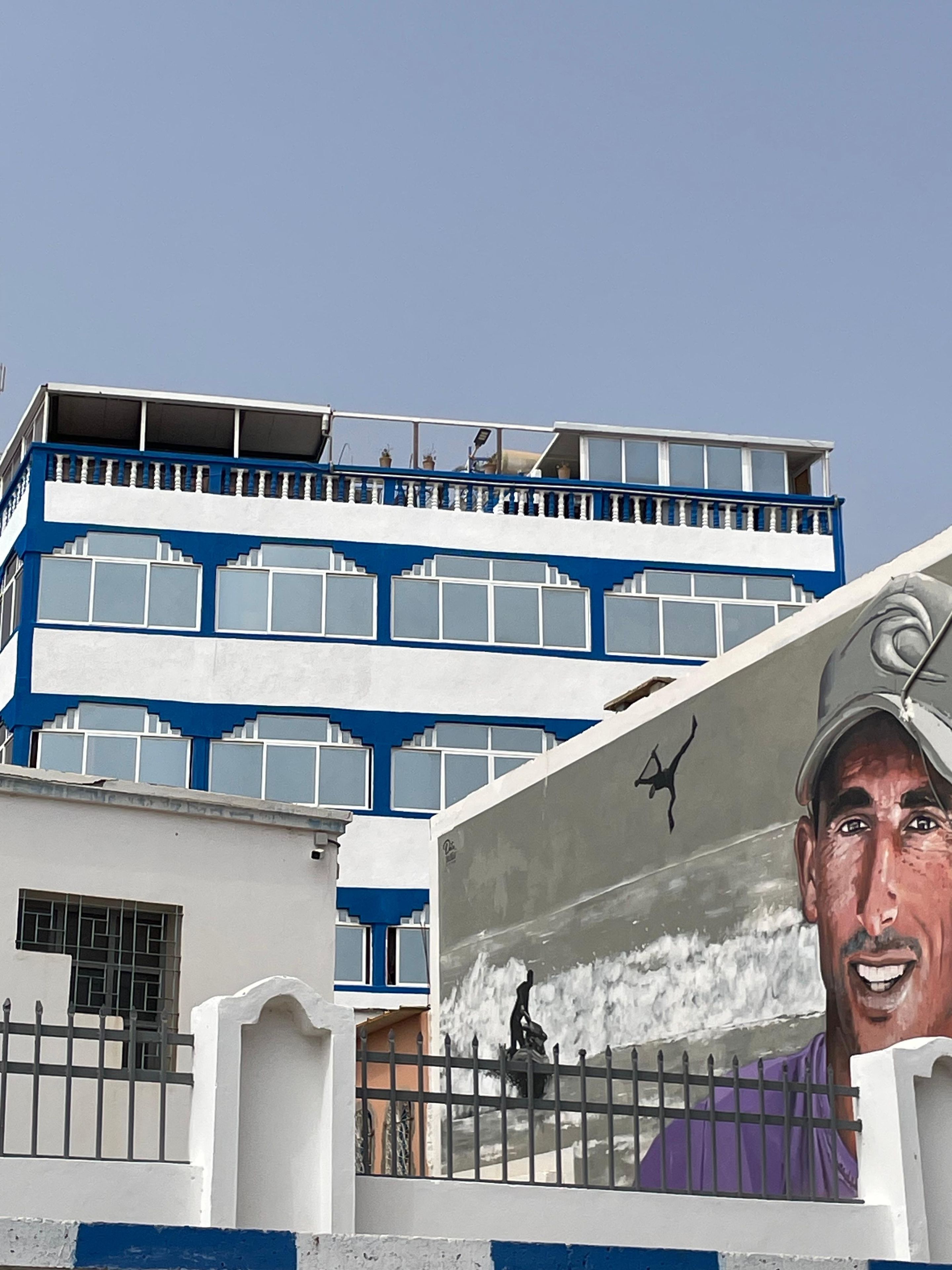 A building with a blue and white facade, a mural with a person, and a fence.