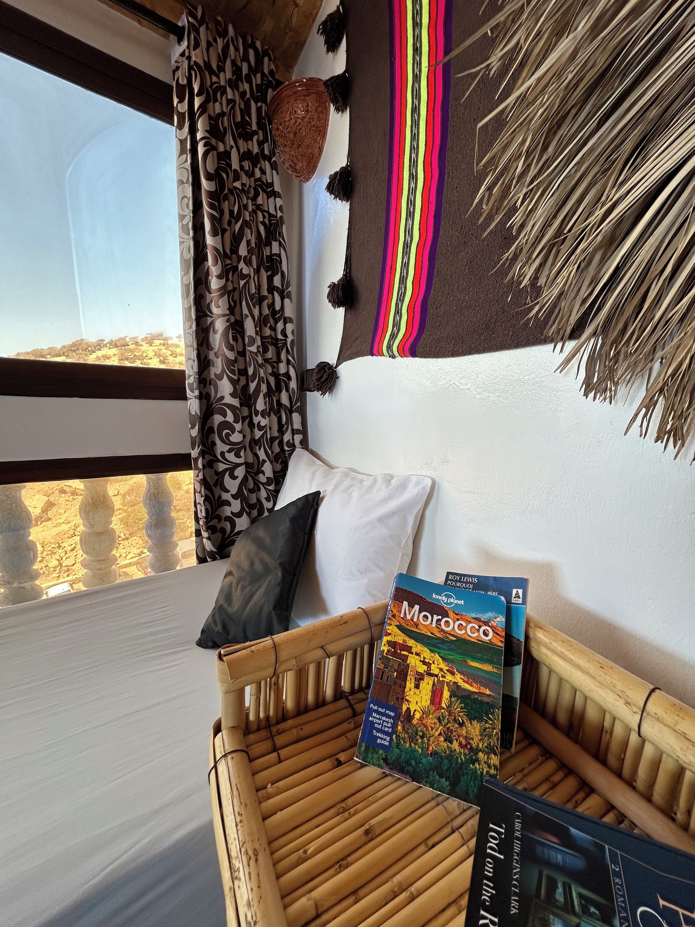 A room with a view of a landscape, a sofa with cushions, a basket, and two travel guides.