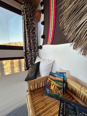 A room with a view of a landscape, a sofa with cushions, a basket, and two travel guides. - AMIDOR HOSTEL (Taghazout)