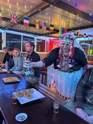 Eine Gruppe von Personen sitzt an einem Tisch mit Speisen und Getränken, eine Person bedient das Essen, und an der Decke hängen Flaggen.