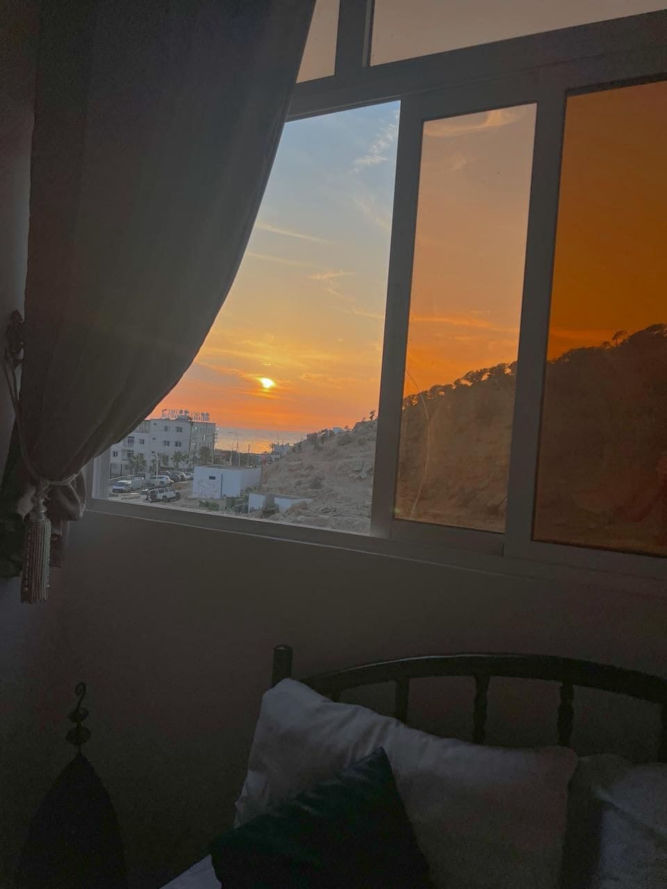 Quarto com cama, janela com vista para o pôr do sol e cortina entreaberta.