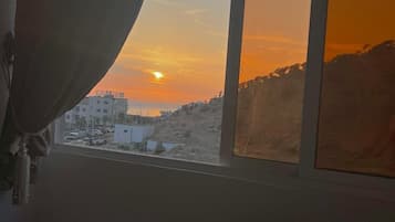 Quarto com cama, janela com vista para o pôr do sol e cortina entreaberta.