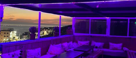 A rooftop terrace with seating and tables, overlooking a cityscape at dusk.