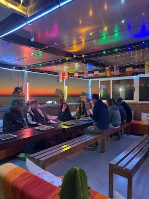 Een restaurant met houten banken en tafels, vlaggen aan de muur en uitzicht op de ondergaande zon.