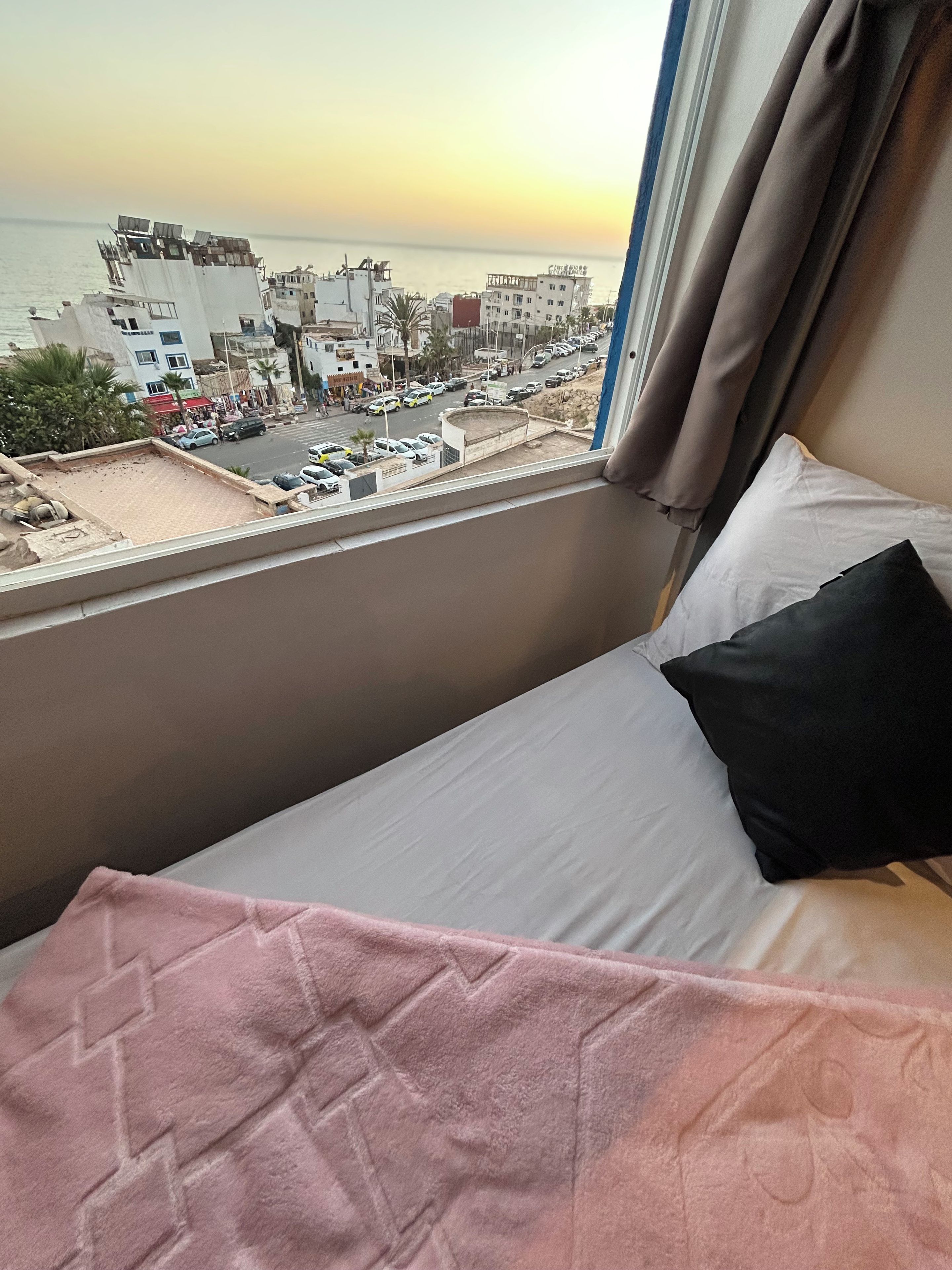 A hotel room with a bed, a window with curtains, and a view of a cityscape and beach.