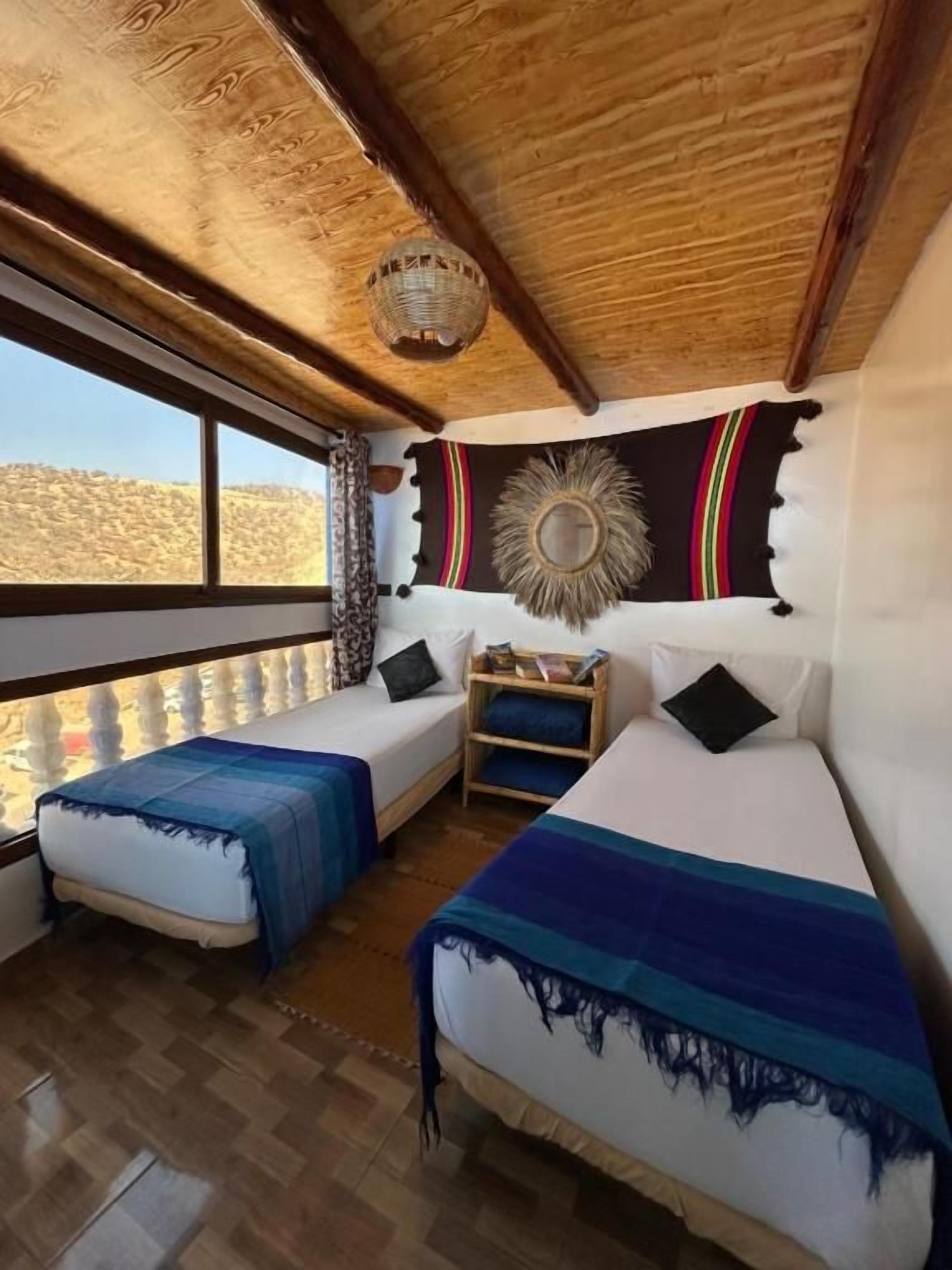 Een kamer met twee bedden, een houten plafond, een groot raam en een decoratief wandkleed.