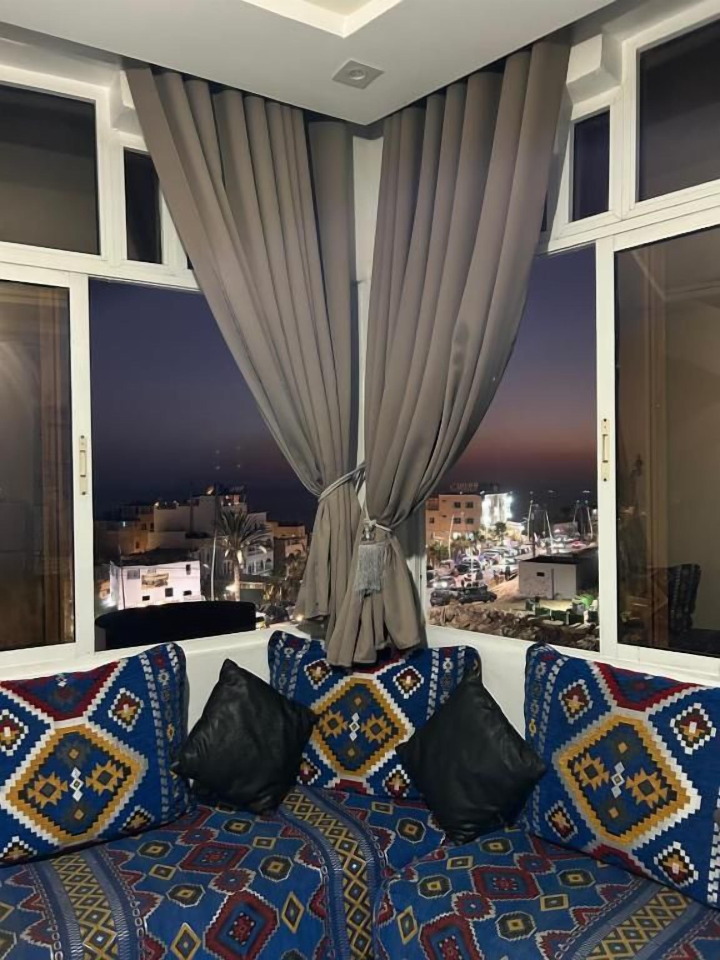 Habitación con vista a un paisaje urbano al atardecer, ventana con cortinas y sofá estampado con cojines.