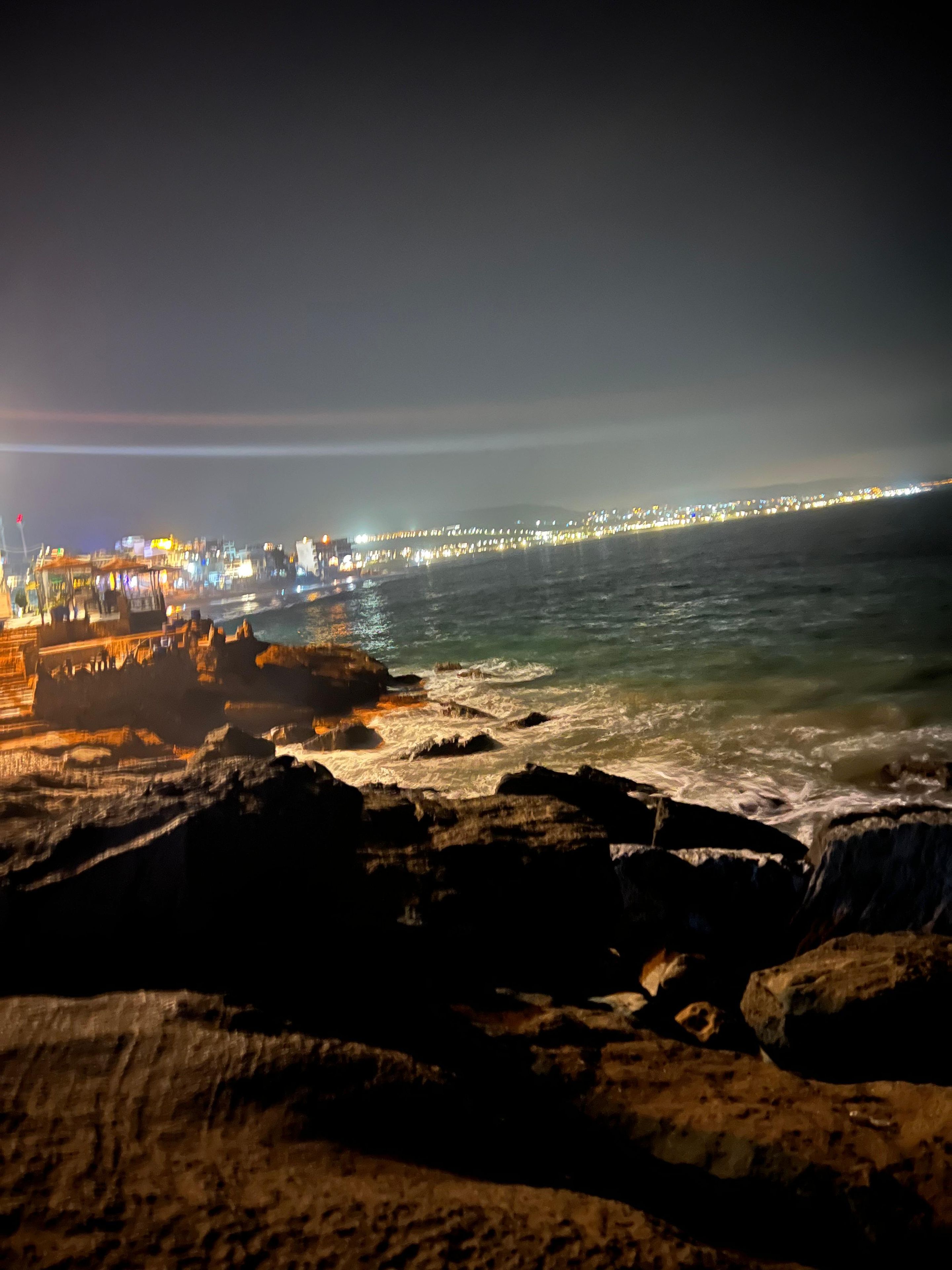 Escena costera nocturna con primer plano rocoso, un muelle que se adentra en el agua y un paisaje urbano con edificios iluminados a lo lejos.
