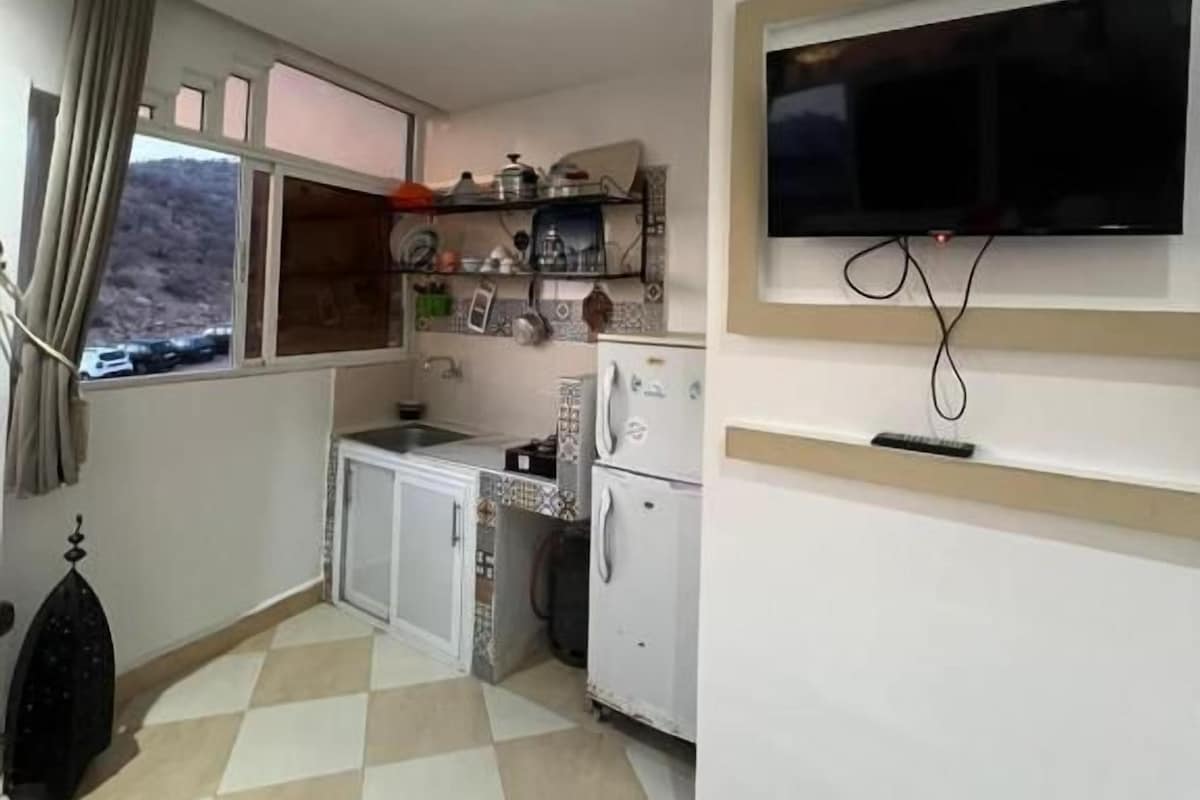 Een compacte keuken met een raam, een flatscreen televisie aan de wand en een kleine koelkast.