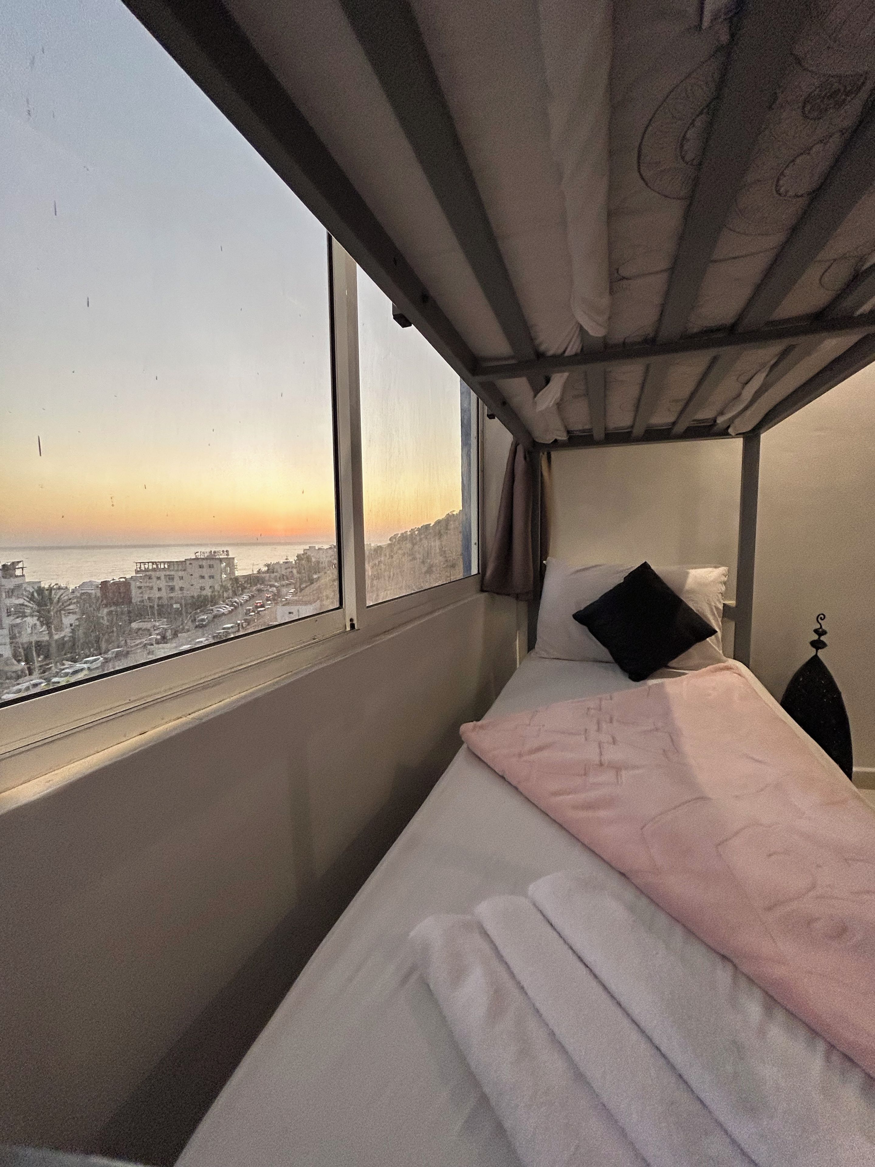 A single bed with a pink blanket and a black pillow, a nightstand, and a window with a cityscape view.