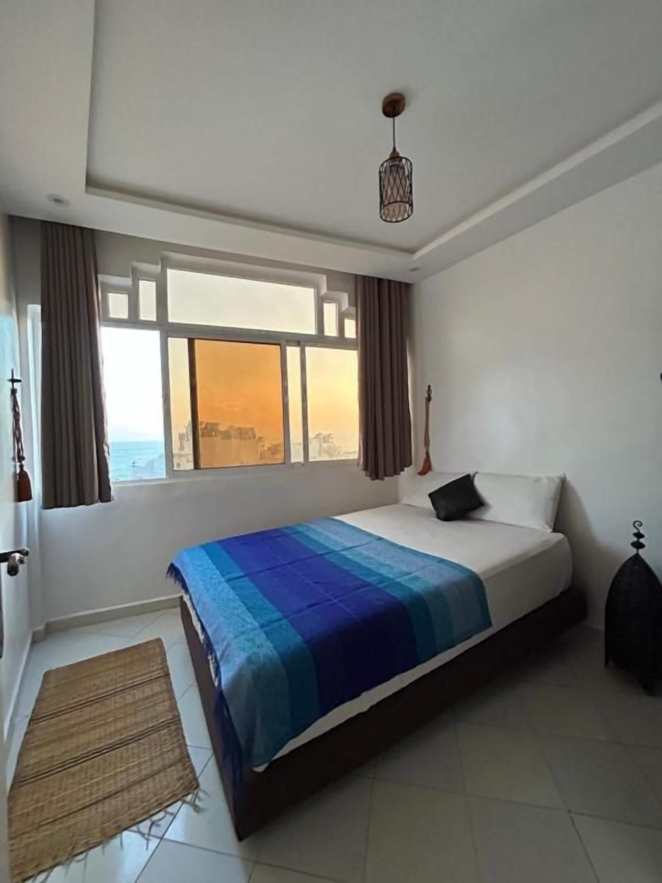 A bedroom with a bed, a window with curtains, a hanging light fixture, and a view of the city.