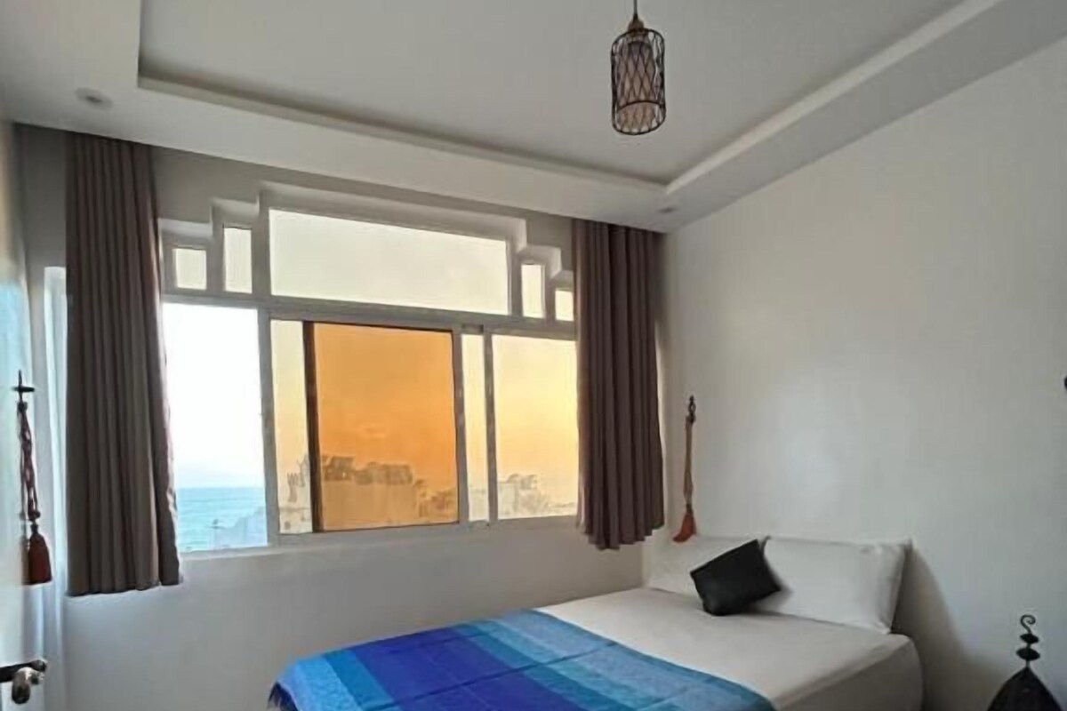 Een slaapkamer met een bed, een raam met gordijnen, een hanglamp en uitzicht op de stad.
