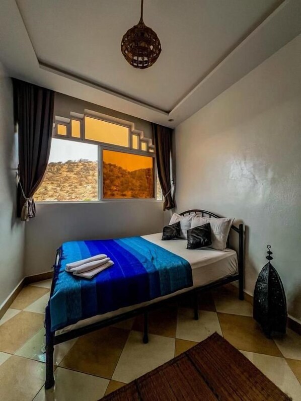 A bedroom with a bed, a window with curtains, a hanging light fixture, and a decorative vase.