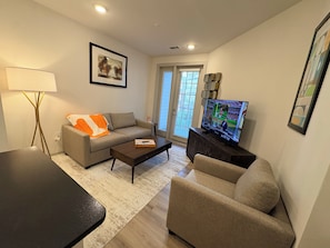 A living room with a sofa, a coffee table, a television, and a lamp. - Riverfront Knoxville Suite - Neyland Stadium Views - Pool + Parking (Knoxville)