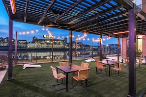 Un espace extérieur aménagé en terrasse, avec des tables et des chaises, une vue sur les gratte-ciel d’une ville au-delà d’une étendue d’eau, et des guirlandes lumineuses.