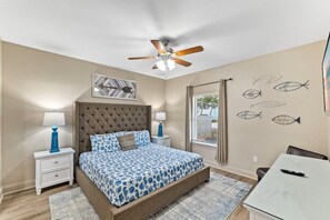 Ein Schlafzimmer mit Bett, Nachttischen, Deckenventilator, Fenster mit Vorhängen und Wandbildern.