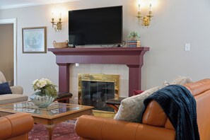 A living room with a flat-screen TV mounted above a fireplace, a sofa, a coffee table, and a bookshelf with books. - Reveille Roundabout: An Aggie Gathering Place 5 min from Kyle Field Sleeps 27 (Bryan)