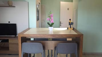 Una pequeña cocina con una mesa de madera, dos taburetes de bar, una orquídea en maceta, un televisor y una puerta con el número 3.