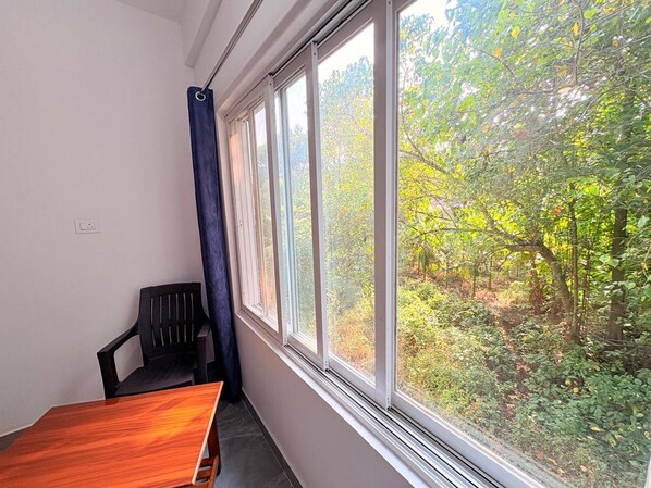 A room with a wooden table and a black chair, a window with a view of a forest, and a power outlet on the wall. - Couple Studio R-8 1st Floor by Mukund Sadan (Arambol)