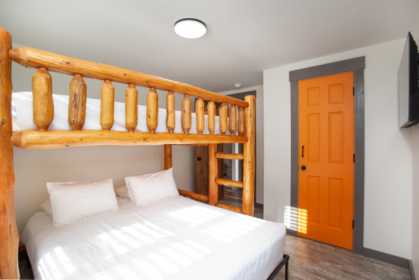 Ein Stockbett mit einer Holzleiter, einer weißen Matratze und Kissen. Es gibt ein Holz-Kopfstück, eine Tür mit orangem Rahmen und eine Deckenleuchte.