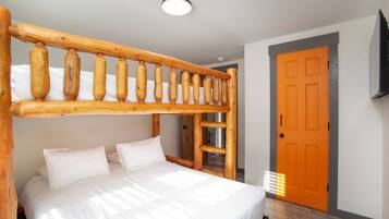 Ein Stockbett mit einer Holzleiter, einer weißen Matratze und Kissen. Es gibt ein Holz-Kopfstück, eine Tür mit orangem Rahmen und eine Deckenleuchte.