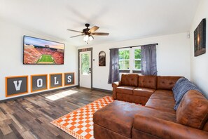 A living room with a brown leather sofa, a ceiling fan, a large framed picture of a stadium, and a rug with a checkered pattern.