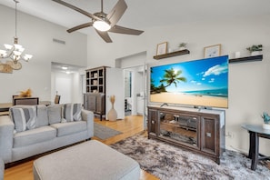 A living room with a flat-screen TV, a ceiling fan, and a view of the beach. - Vista Verde East 207 (St. Petersburg)