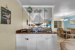 A kitchen with a central island, a dining area with a table and chairs, and a decorative sign on the wall. - Barefoot Beach Resort B201 (Indian Shores)