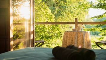A room with a bed, a small table with a floral tablecloth, and a view of green trees through a window.